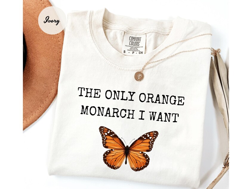 Pode incluir: Camiseta cor marfim com o texto "THE ONLY ORANGE MONARCH I WANT" acima de um gr&aacute;fico detalhado de uma borboleta laranja e preta. Um colar de ouro com um pingente est&aacute; drapeado sobre a camiseta.