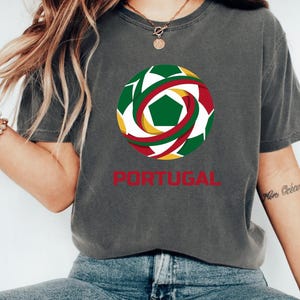 May include: Dark grey t-shirt featuring a soccer ball design with green, red, yellow, and white accents. The word "PORTUGAL" is printed in red below the graphic. The shirt is a casual style.