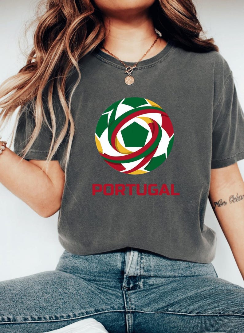 May include: Dark grey t-shirt featuring a soccer ball design with green, red, yellow, and white accents. The word "PORTUGAL" is printed in red below the graphic. The shirt is a casual style.