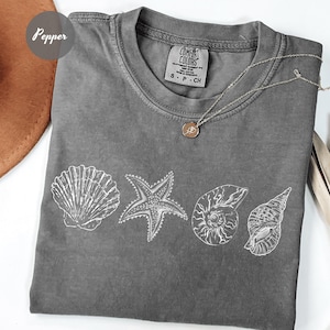 May include: A gray Comfort Colors t-shirt featuring white line drawings of seashells and a starfish. A silver necklace with a small pendant rests on the shirt. The shirt is a size small.