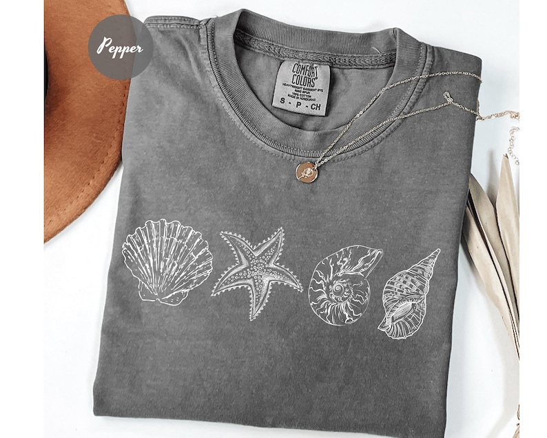 May include: A gray Comfort Colors t-shirt featuring white line drawings of seashells and a starfish. A silver necklace with a small pendant rests on the shirt. The shirt is a size small.