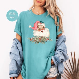 May include: A turquoise t-shirt featuring a vintage Santa Claus graphic. Santa wears a pink hat and is surrounded by greenery. The text "Chalky Mint" is in a circle on the upper left. The shirt has rolled-up sleeves.