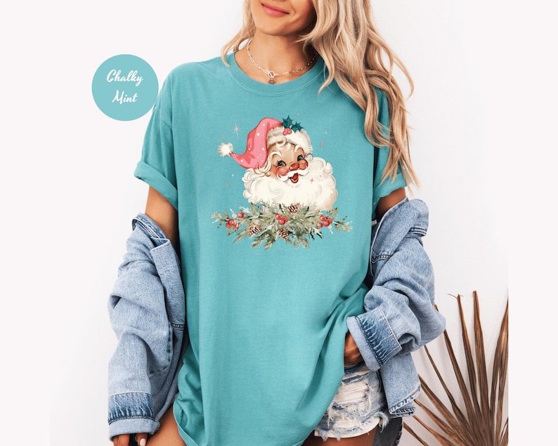 May include: A turquoise t-shirt featuring a vintage Santa Claus graphic. Santa wears a pink hat and is surrounded by greenery. The text "Chalky Mint" is in a circle on the upper left. The shirt has rolled-up sleeves.