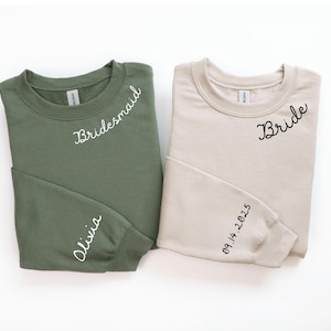 May include: Two crewneck sweatshirts, one olive green and one beige. The olive green sweatshirt has "Bridesmaid" and "Olivia" in white script. The beige sweatshirt has "Bride" and "09.14.2025" in black script.