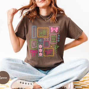 May include: Espresso-colored t-shirt featuring a colorful graphic of postage stamp-like designs with flowers, bows, and text. The shirt is made of a soft material and has a relaxed fit. The model is wearing light blue jeans and white sneakers.