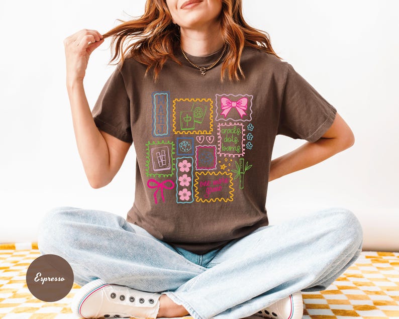 May include: Espresso-colored t-shirt featuring a colorful graphic of postage stamp-like designs with flowers, bows, and text. The shirt is made of a soft material and has a relaxed fit. The model is wearing light blue jeans and white sneakers.