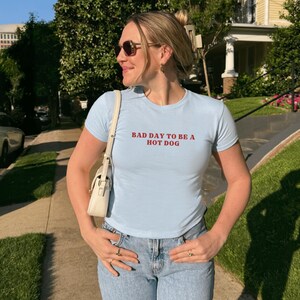 Puede incluir: Camiseta azul claro de cuello redondo con el texto rojo "BAD DAY TO BE A HOT DOG". La camiseta se combina con vaqueros de mezclilla de lavado claro. Un bolso blanco se lleva al hombro.