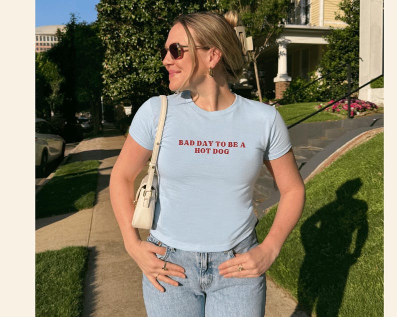 Puede incluir: Camiseta azul claro de cuello redondo con el texto rojo "BAD DAY TO BE A HOT DOG". La camiseta se combina con vaqueros de mezclilla de lavado claro. Un bolso blanco se lleva al hombro.