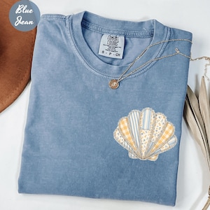 May include: A blue Comfort Colors t-shirt with a seashell applique in various patterns of yellow, white, and blue. A gold necklace with a pendant rests on the shirt. The shirt is folded on a white surface.