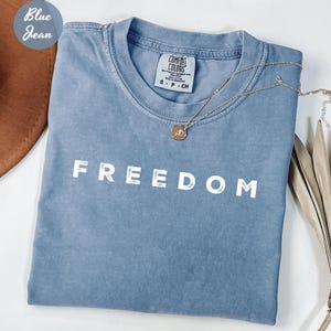 May include: A blue jean-colored t-shirt with the word "FREEDOM" printed in white block letters. A gold necklace with a pendant rests on the shirt. The shirt is folded and placed next to a brown hat.