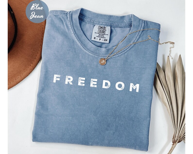 May include: A blue jean-colored t-shirt with the word "FREEDOM" printed in white block letters. A gold necklace with a pendant rests on the shirt. The shirt is folded and placed next to a brown hat.