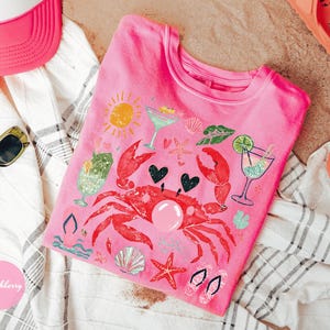 May include: Pink t-shirt with a crab graphic, cocktails, and beach-themed illustrations. The shirt is surrounded by a pink visor, sunglasses, and orange sandals, suggesting a summer theme. The design includes hearts and a sun.