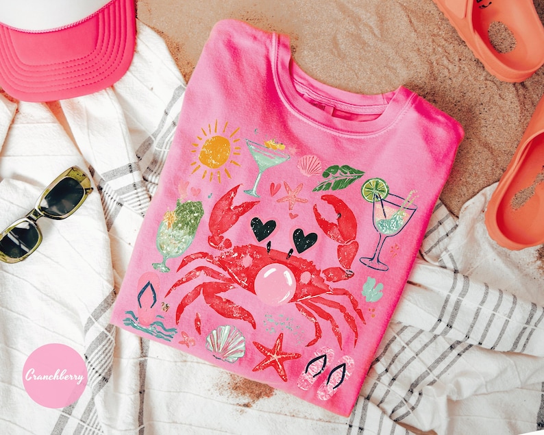 May include: Pink t-shirt with a crab graphic, cocktails, and beach-themed illustrations. The shirt is surrounded by a pink visor, sunglasses, and orange sandals, suggesting a summer theme. The design includes hearts and a sun.