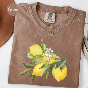 May include: Espresso-colored t-shirt with a watercolor illustration of lemons, green leaves, and white flowers. The shirt has a crew neck and short sleeves. A silver necklace with a small pendant is draped on the shirt.