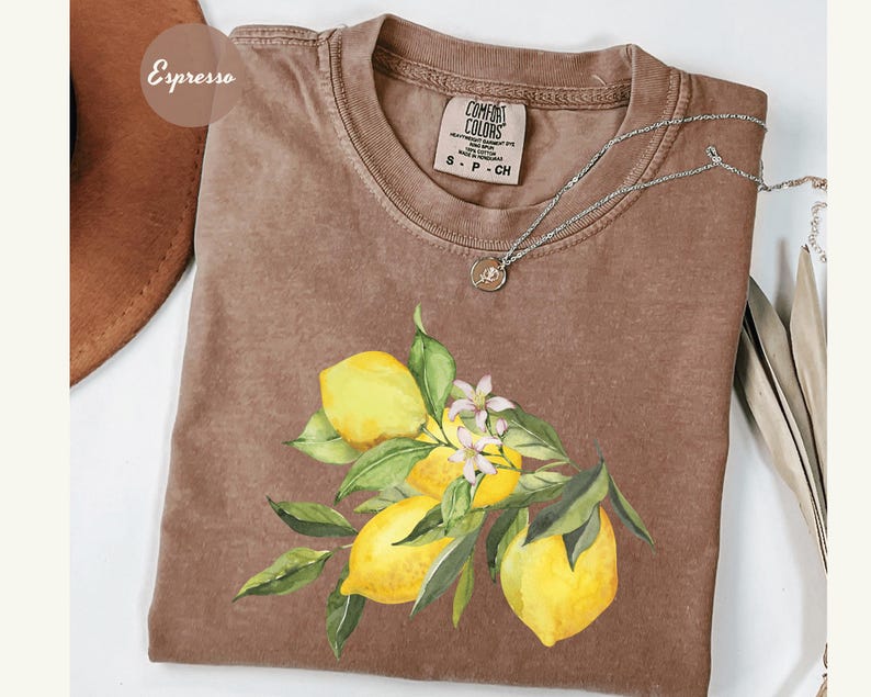 May include: Espresso-colored t-shirt with a watercolor illustration of lemons, green leaves, and white flowers. The shirt has a crew neck and short sleeves. A silver necklace with a small pendant is draped on the shirt.