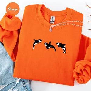 May include: Orange sweatshirt featuring three black and white orca whale graphics. The sweatshirt has a crew neck and long sleeves. A silver necklace with a sun pendant is draped across the top. The word "Orange" is printed on a circular sticker.