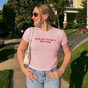 Puede incluir: Camiseta rosa claro con el texto rojo "BAD DAY TO BE A HOT DOG". La camiseta es de manga corta y se lleva con vaqueros azules. Un bolso blanco se lleva al hombro.