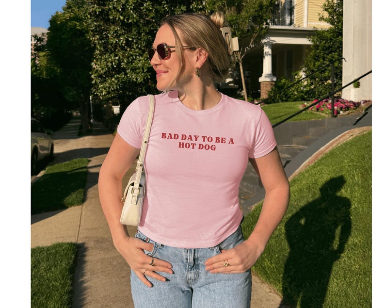 Puede incluir: Camiseta rosa claro con el texto rojo "BAD DAY TO BE A HOT DOG". La camiseta es de manga corta y se lleva con vaqueros azules. Un bolso blanco se lleva al hombro.