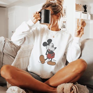May include: White sweatshirt featuring a classic Mickey Mouse graphic in black, red, and tan. The sweatshirt has a crew neck and long sleeves. A black mug is held up near the face. The person is sitting on a couch.