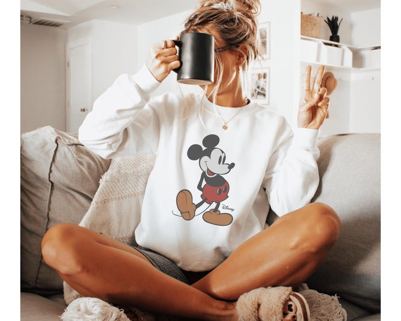 May include: White sweatshirt featuring a classic Mickey Mouse graphic in black, red, and tan. The sweatshirt has a crew neck and long sleeves. A black mug is held up near the face. The person is sitting on a couch.
