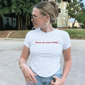 May include: White t-shirt with the red text "Show me your books." The shirt is short-sleeved and has a crew neck. The person in the image is wearing gold-coloured jewellery and sunglasses. The background shows a residential street.