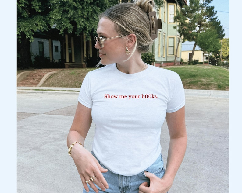 May include: White t-shirt with the red text "Show me your books." The shirt is short-sleeved and has a crew neck. The person in the image is wearing gold-coloured jewellery and sunglasses. The background shows a residential street.