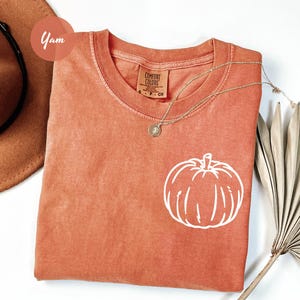 May include: A coral-coloured t-shirt with a white pumpkin graphic. The shirt has a crew neck and short sleeves. A gold necklace with a pendant is draped across the shirt. A brown hat and dried palm leaves are also in the image.