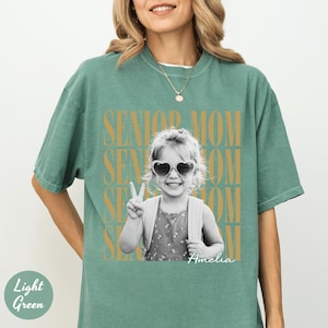 May include: A light green short-sleeve t-shirt featuring a black and white photograph of a child wearing heart-shaped sunglasses and making a peace sign. The text SENIOR MOM is repeated in gold behind the photo, with the name Amelia in script.