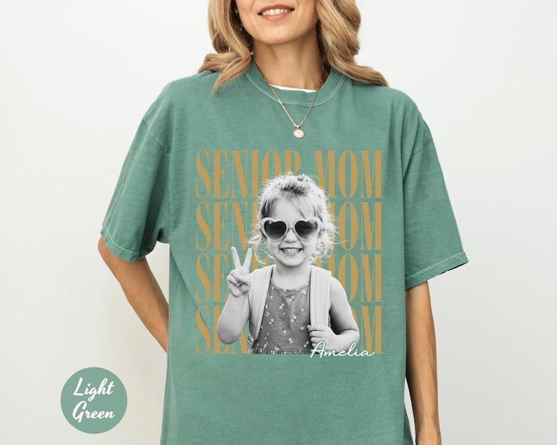 May include: A light green short-sleeve t-shirt featuring a black and white photograph of a child wearing heart-shaped sunglasses and making a peace sign. The text SENIOR MOM is repeated in gold behind the photo, with the name Amelia in script.