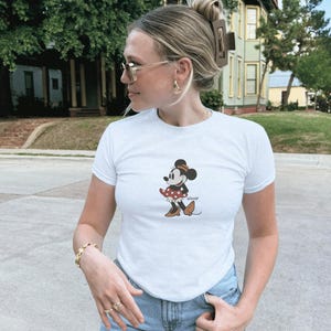 Pu&ograve; includere: T-shirt bianco con grafica di Minnie Mouse. Il personaggio dei cartoni animati &egrave; raffigurato in una posa classica, con un vestito a pois rossi e scarpe marroni. La maglietta ha maniche corte e girocollo.