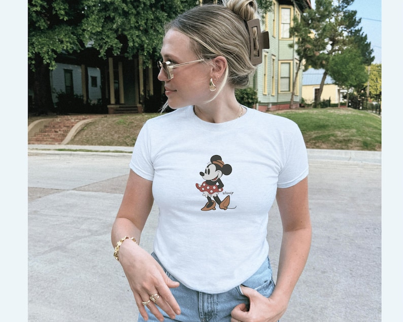 Pu&ograve; includere: T-shirt bianco con grafica di Minnie Mouse. Il personaggio dei cartoni animati &egrave; raffigurato in una posa classica, con un vestito a pois rossi e scarpe marroni. La maglietta ha maniche corte e girocollo.
