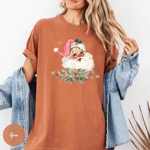 May include: A rust-colored t-shirt featuring a Santa Claus graphic. The Santa illustration has a pink hat, white beard, and is surrounded by a festive wreath. The shirt has short sleeves and a relaxed fit, perfect for the holiday season.