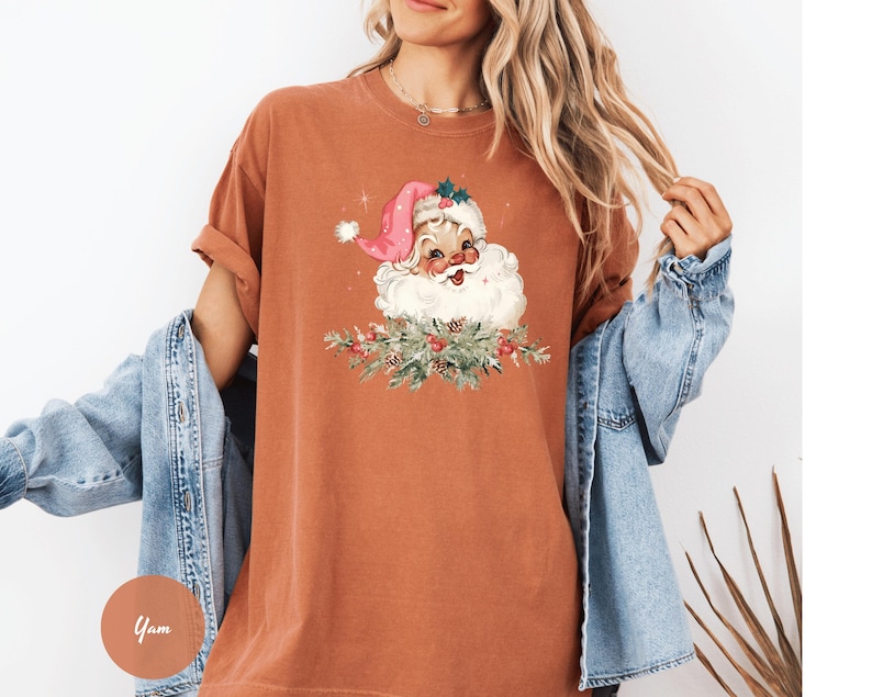 May include: A rust-colored t-shirt featuring a Santa Claus graphic. The Santa illustration has a pink hat, white beard, and is surrounded by a festive wreath. The shirt has short sleeves and a relaxed fit, perfect for the holiday season.