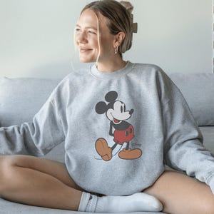 May include: A grey sweatshirt with a Mickey Mouse design. The cartoon character is shown in his classic pose, wearing red shorts, brown shoes, and white gloves. The Disney logo is visible on the lower right.
