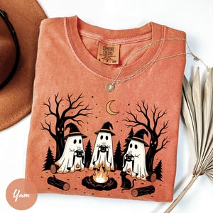 Peut inclure: Un t-shirt orange brûlé avec un motif de trois fantômes portant des chapeaux de sorcière assis autour d'un feu de camp dans une forêt. Les fantômes tiennent des tasses et un chat noir est assis à proximité. Étiquette Comfort Colors au col.