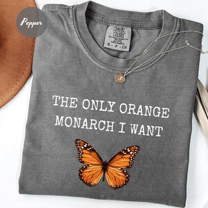 Pode incluir: Camiseta cinza Comfort Colors com o texto "THE ONLY ORANGE MONARCH I WANT" e um gr&aacute;fico de borboleta monarca. Um colar de prata com um pingente est&aacute; na camisa.