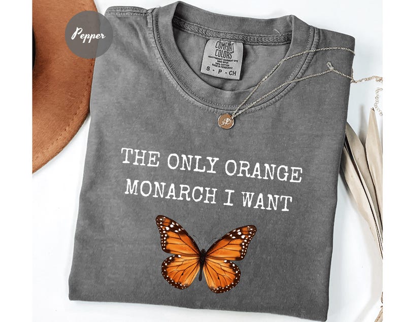 Pode incluir: Camiseta cinza Comfort Colors com o texto "THE ONLY ORANGE MONARCH I WANT" e um gr&aacute;fico de borboleta monarca. Um colar de prata com um pingente est&aacute; na camisa.