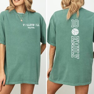 May include: A sage green t-shirt with the text "VOLLEYBALL mom" on the front. The back of the shirt features the number 10, a volleyball graphic, and the name "EMILY" in white. The shirt has short sleeves and a relaxed fit.