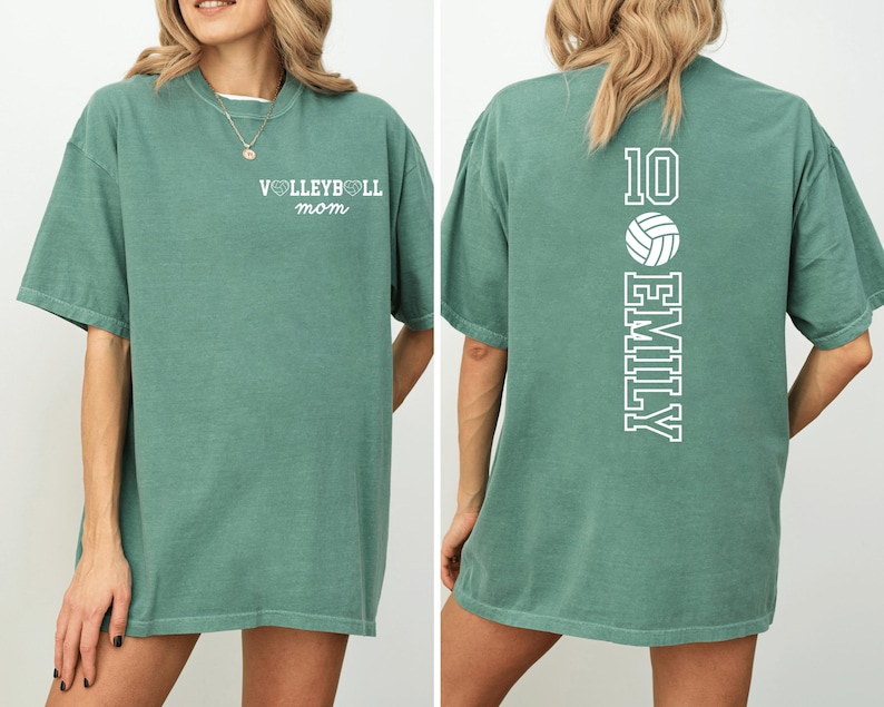 May include: A sage green t-shirt with the text "VOLLEYBALL mom" on the front. The back of the shirt features the number 10, a volleyball graphic, and the name "EMILY" in white. The shirt has short sleeves and a relaxed fit.