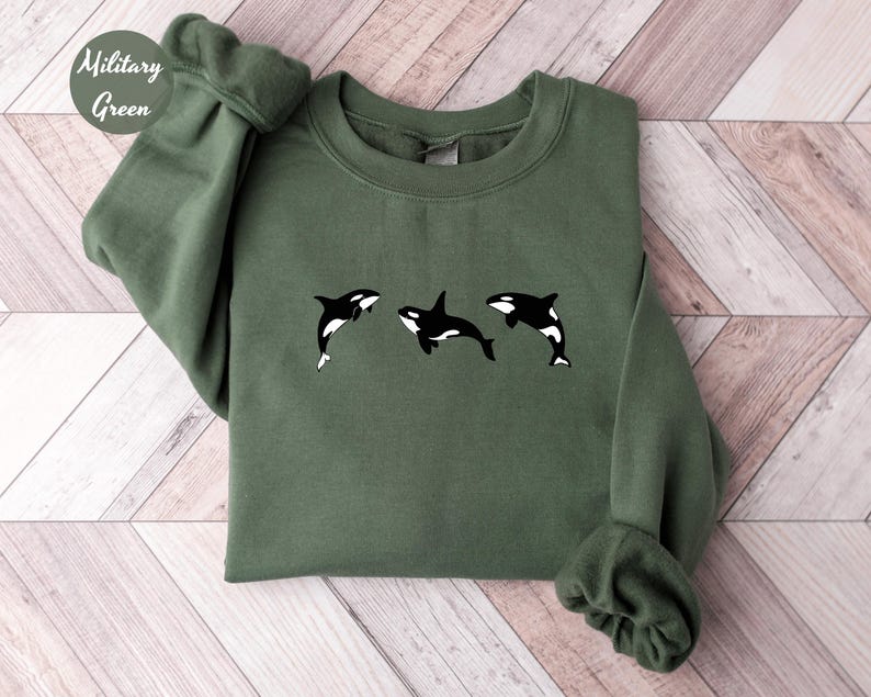 May include: Military green sweatshirt featuring three black and white orca whale designs. The sweatshirt has a crew neck and long sleeves. The text "Military Green" is in a circle in the upper left corner.