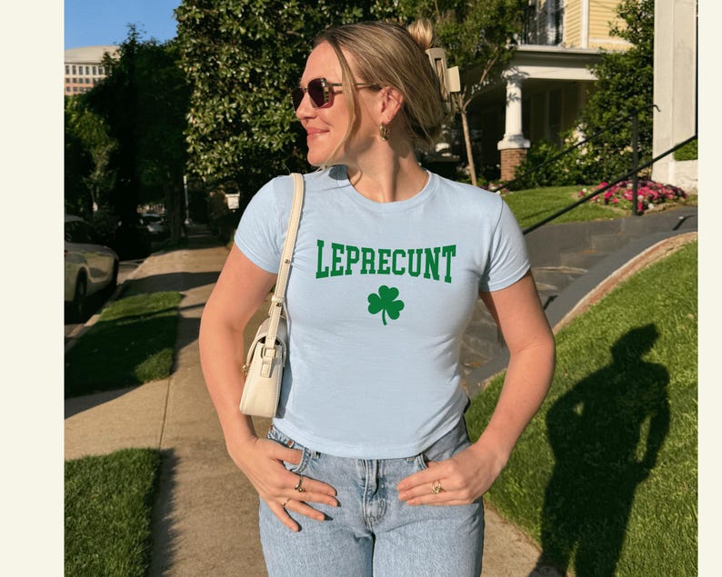 May include: A pale blue t-shirt with the word "LEPRECUNT" in green block letters and a green shamrock graphic. The shirt is worn with light blue jeans.