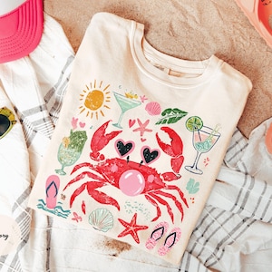 May include: Ivory-colored t-shirt with a colorful beach-themed graphic. The design includes a red crab wearing heart-shaped sunglasses, cocktails, seashells, and a sun. The shirt is laid on a striped towel with sunglasses, a pink hat, and sandals.