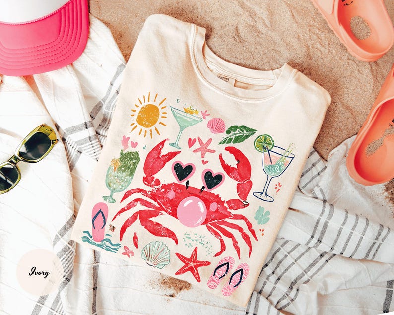 May include: Ivory-colored t-shirt with a colorful beach-themed graphic. The design includes a red crab wearing heart-shaped sunglasses, cocktails, seashells, and a sun. The shirt is laid on a striped towel with sunglasses, a pink hat, and sandals.
