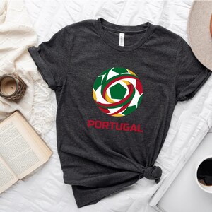 May include: Dark gray t-shirt featuring a colorful soccer ball design with green, red, and yellow accents. The word "PORTUGAL" is printed in red below the graphic. The shirt is knotted at the waist.