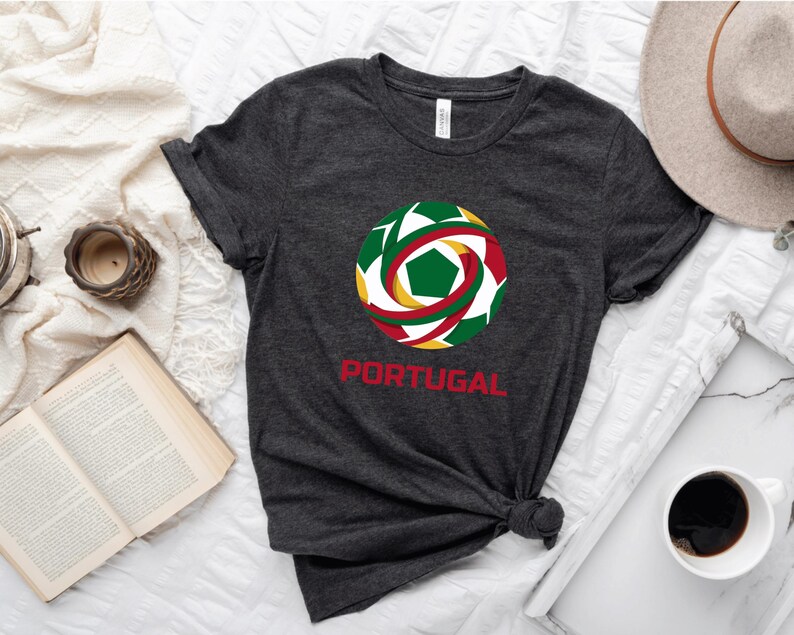May include: Dark gray t-shirt featuring a colorful soccer ball design with green, red, and yellow accents. The word "PORTUGAL" is printed in red below the graphic. The shirt is knotted at the waist.