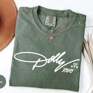 May include: A moss green short-sleeve t-shirt featuring the white printed text Dolly xoxo with three small heart outlines. The shirt has a crew neckline and a Comfort Colors brand label on the inside collar indicating a size small.