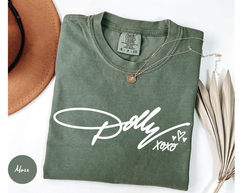 May include: A moss green short-sleeve t-shirt featuring the white printed text Dolly xoxo with three small heart outlines. The shirt has a crew neckline and a Comfort Colors brand label on the inside collar indicating a size small.