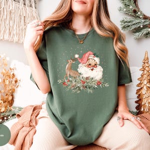 May include: A moss green t-shirt featuring a vintage Christmas graphic. The graphic includes Santa Claus with a pink hat, a reindeer, and festive greenery. The shirt is a relaxed fit, perfect for the holiday season.