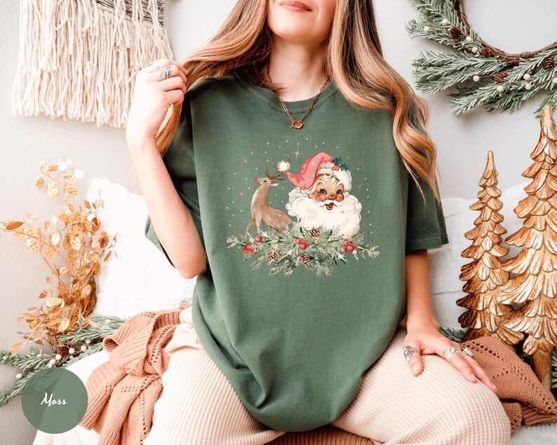 May include: A moss green t-shirt featuring a vintage Christmas graphic. The graphic includes Santa Claus with a pink hat, a reindeer, and festive greenery. The shirt is a relaxed fit, perfect for the holiday season.