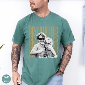 May include: Light green t-shirt with the text "BEST DAD EVER" in gold lettering. A black and white photo of two children wearing sunglasses is printed on the shirt, with the names "Sam & Olivia" below.